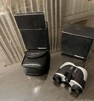 Two Tasco small binoculars placed on table with their respective black zippered carrying cases and original black Tasco boxes.