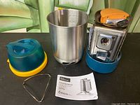 Photo of disassembled BioLite Camp Stove showing stove unit with attached fan and fuel holder, stainless steel pot, vented lid, metal stand, and instruction manual on table.