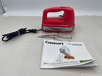 Red Cuisinart hand mixer with black speed control slider, chrome detailing, two stainless steel beaters, and instruction manual laid out on white background.