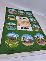 Full view of green bordered linen tea towel featuring colored illustrations of Cotswold Country landmarks including Cheltenham, Stow on the Wold, and Chipping Campden. Printed 100% linen by Ulster.