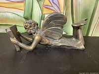 Side view showing full profile of metal angel figurine lying down reading a book.