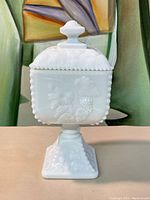 Front view of vintage milk glass covered dish showing square shape, beaded edge detailing, lid with finial, and embossed grape and leaf motifs.