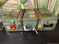 Front view of vintage green suitcase with leather straps and multiple travel stickers including circular and rectangular shapes.