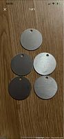 Five round metal tags displayed on a wooden surface, showing the front side and hole at the top edge.