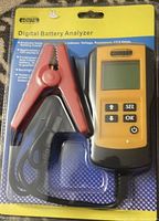Front view of the digital battery analyzer in sealed packaging showing yellow handheld device with LCD screen and control buttons, connected to red and black alligator clips via a coiled cable.