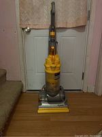 Full view of the upright Dyson vacuum cleaner showing its yellow and gray body, standing upright against a white door indoors.