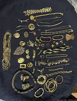 Photo showing an assortment of gold-toned costume jewellery laid out on black fabric including necklaces, bracelets, brooches, rings and earrings.