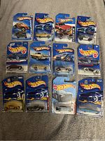 Hot Wheels lot of 12 cars in original blister card packaging on carpet background, showing various models and colors.