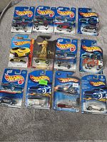 Photo showing 11 Hot Wheels diecast cars in blue packaging arranged in rows on a carpeted floor.
