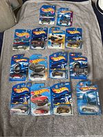 Fourteen Hot Wheels die-cast cars in original blister packaging laid out on a grey fabric background, showing a variety of models and colors.