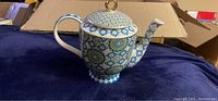 Photo showing the teapot with intricate Isfara geometric pattern in blue and gold, placed on a dark fabric surface.