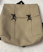 Front view of beige RAINS MSN backpack bag showing flap top and black buckle clasps