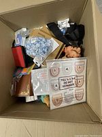 Open cardboard box containing various stationery items such as an envelope set with rainbow patterns, packaged cards, gift bags, fabric pieces, and a small plush toy.