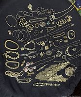 Top-down image of assorted silver-toned costume jewellery spread out on dark fabric including various bracelets, necklaces, rings, earrings, and brooches.