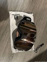 Photo of Solomon snowboarding goggles with reflective lens lying on white Solomon branded protective bag on wood floor, showing front of goggles.