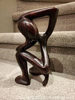 Front view of dark wood abstract human figure sculpture sitting on carpeted stairs.