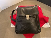 Front view of black leather backpack-style bag with flap and metal clasp closure on red and white fabric.