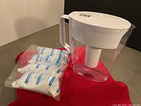Clear plastic Brita water filter jug with white lid and handle on gray surface with a red cloth, showing 4 new sealed filter cartridges.