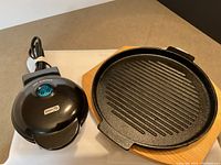 Cast iron grill pan and DASH mini waffle maker side by side on counter showing grill pan wooden trivet and waffle maker cord.