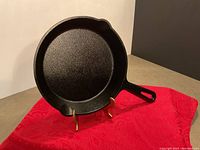 Front view of black pre-seasoned cast iron skillet with handle and small pouring spouts on both sides. The pan is standing on a display stand over a red cloth.