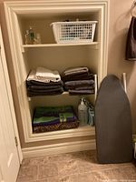 Photo showing bathroom shelves stocked with folded bath towels, beach towels, toiletries in bottles and containers, some items in white plastic basket, and a dark gray tabletop ironing board leaning against the wall.