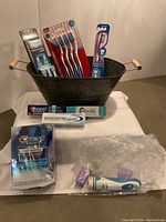 Photo showing Oral-B electric toothbrush, manual toothbrushes in packaging, Sensodyne and Aquafresh toothpaste boxes, and Crest 3D White strips pack on white surface within black bucket.