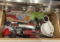Photo showing the drawer with various kitchen utensils laid out, revealing ladles, graters, peelers, scissors, whisks, and slicers in a somewhat organized manner.