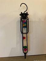 Full view of the hanging thermometer with black metal frame and hook, showing the sealed glass cylinder containing several colorful floating glass spheres.