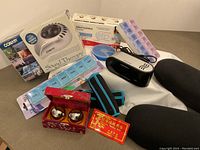 View of all health-related items including therapy machine box, pill organizer, black support cushions, Chinese stress balls in red box, digital clock, and black/blue straps.