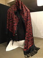 Scarf draped over a mannequin showing full length and pattern on black background with red ornamental designs and fringed ends.