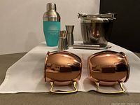 Photo showing the copper Moscow Mule mugs front and center with brass handles, cocktail shaker with a teal wrap, stainless steel ice bucket with tongs, and two cocktail jiggers arranged behind.