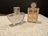 Two perfume bottles side by side on a white doily. Left is Nautica Bermuda Blue with light liquid, right is Burberry with amber liquid and plaid design on the bottle.
