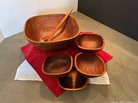 Full view of the five-piece teak bowl set and serving spoon placed on fabric