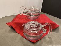 Two identical clear glass Pyrex tea pots side by side on a red cloth, showing overall condition and shape.