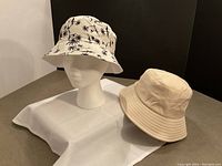 Two bucket hats on white cloth, one beige and one off-white with palm tree print, displayed on a head mannequin and lying flat.
