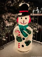 Snowman blow mold at night on snow-covered outdoor porch, lit from within showing its translucent body and painted details.
