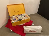 Yellow vintage two-level hard plastic sewing box open showing multiple compartments filled with spools of thread and sewing accessories, red cloth under the box, white Handy Stitch bag beside it.