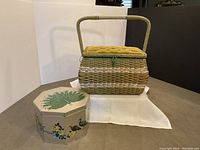 Rectangular woven sewing basket with mustard yellow tufted velvet lid and octagonal storage tin with floral and peacock painting on cream background, displayed on table.