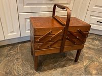 Closed wooden accordion-style sewing box showing expanded sides, carrying handle, and legs on tiled floor.