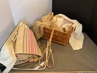 Photo displaying the folded vintage cream and pink wool blanket next to two textured handmade throws, a wicker basket containing throws, and a wicker rug beater placed on a surface.