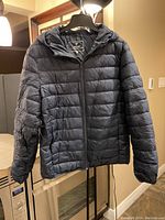 Front view of navy blue puffer jacket with attached hood hanging on a mannequin in indoor setting, showing full zipper and horizontal stitching.
