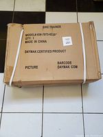 Closed cardboard shipping box with printed product details stating model KW-7073-0220, 'Bike Trainer', brand Daymak, Made in China.