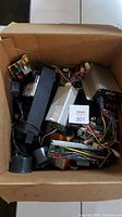 A box filled with various untested electrical parts including power supplies, meters, circuit boards, and wiring.