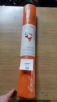 Rolled up orange Jogi yoga mat with original packaging band, held upright on wooden floor.