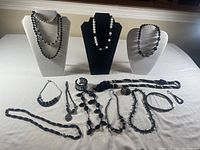 Black costume jewellery pieces displayed on busts and laid on white cloth including necklaces, bracelets, and pins.