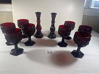 Set of six burgundy glass wine goblets and two matching burgundy glass candlesticks on a flat surface with neutral background.