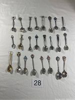 Flat lay photo of 23 decorative collectors teaspoons arranged in 3 rows on white surface with an inventory number '28' marker at bottom center.