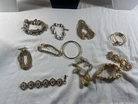 Photo showing the 11 gold-tone costume bracelets arranged on white surface; various designs including chain, mesh, bangle, and decorated fabric insets visible. Some wear and damage visible.