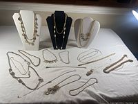 Full display of about 20 pieces of costume jewellery on white cloth with three necklace busts showing multiple gold tone necklaces with various designs including coin charms, linked chains, and decorative elements.