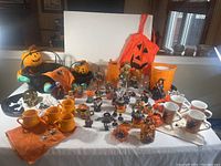 Photo showing arrangement of Halloween-themed mugs, baskets with pumpkin plushes, orange towels, decorative plates, and various small Halloween figurines on a white cloth-covered table